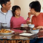 An Asian family, an adult male and female are seated around a table eating a meal with a young female standing in between the adults.